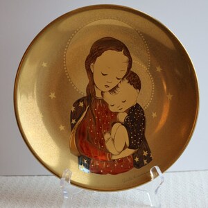 Op de afbeelding: Een gouden keramische plaat met een geschilderd beeld van een moeder en kind. De moeder houdt het kind in haar armen en ze kijken allebei naar beneden. De plaat heeft een gouden achtergrond met een witte halo rond de moeder en het kind. De plaat staat op een wit oppervlak.
