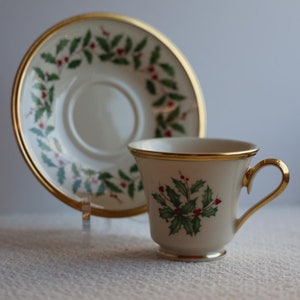 May include: A white teacup and saucer with a gold rim and a green and red holly design.