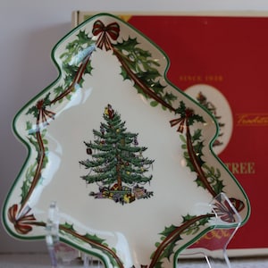 Puede incluir: Un plato de servir de cerámica blanca con forma de árbol de Navidad con un borde verde y un diseño de cinta verde y roja. El plato presenta una imagen pintada de un árbol de Navidad con adornos y regalos. El plato está sobre una superficie blanca con un soporte de acrílico transparente.