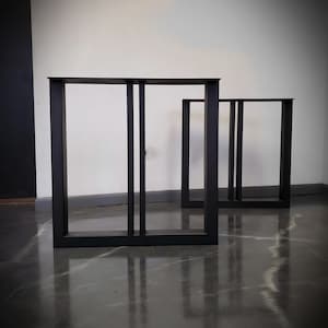 May include: Two black metal table legs with a rectangular frame design. The legs are standing on a gray concrete floor.