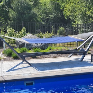 May include: A blue hammock with a black metal frame is set up near a swimming pool. The hammock is hanging between two posts and is made of a woven fabric. The pool is surrounded by a stone patio and there is a small fountain in the foreground.