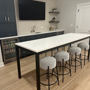 May include: A white countertop bar table with black metal legs and five gray upholstered bar stools. The table is in a basement with a dark blue cabinet and a mini fridge.