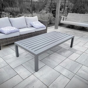 May include: A gray rectangular coffee table with a slatted top sits on a gray patio. The table is made of metal and has a modern design.
