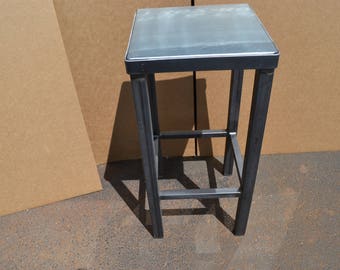May include: A black metal stool with a square top and a gray top. The stool has a simple design with a single crossbar for support.