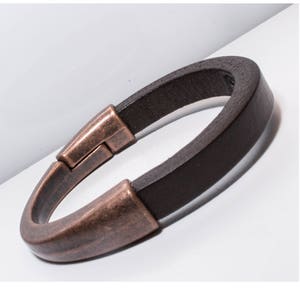 Op de afbeelding: Een bruin leren armband met een koperkleurige sluiting. De armband is gemaakt van een enkel stuk leer dat is gevouwen en met de sluiting is vastgemaakt.