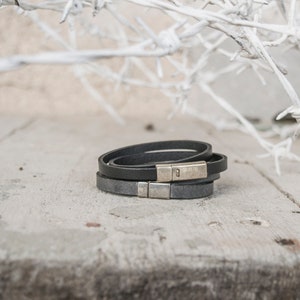 Op de afbeelding: Drie zwarte leren armbanden met zilveren magneetsluitingen. De armbanden liggen op een lichtgrijs houten oppervlak.