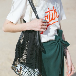 Op de afbeelding: Een vrouw draagt een wit T-shirt met het woord "batik" in rood gedrukt op de voorkant. Ze draagt een zwarte mesh-tas met een leren handvat. De tas is gevuld met een item met een patroon. Ze draagt ook een donkergroene wikkelrok.