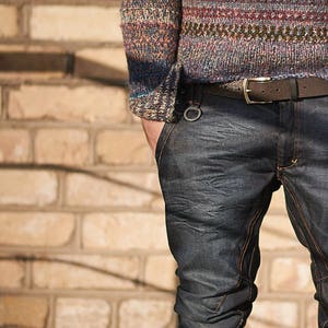 Op de afbeelding: Een persoon draagt een blauw-bruine gestreepte trui met een bruine leren riem en een donkerblauwe jeans.