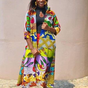 May include: A woman wearing a long, colourful patterned cardigan with a black shirt underneath. She is standing in front of a light brown wall and smiling at the camera. The cardigan is made of different fabrics and has a patchwork design. The woman is wearing black high heels.