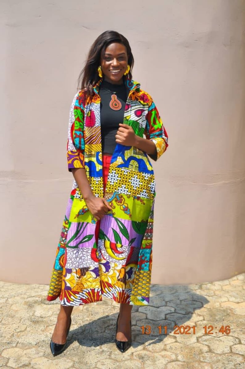 May include: A woman wearing a long, colourful patterned cardigan with a black shirt underneath. She is standing in front of a light brown wall and smiling at the camera. The cardigan is made of different fabrics and has a patchwork design. The woman is wearing black high heels.