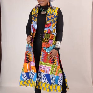 Puede incluir: Un chaleco largo, colorido y estampado con cuello blanco y abertura frontal. El chaleco está hecho de un patchwork de diferentes telas con diseños geométricos y florales en tonos rojos, naranjas, amarillos, azules, verdes y morados. El chaleco se lleva sobre una camisa negra de manga larga y pantalones negros.