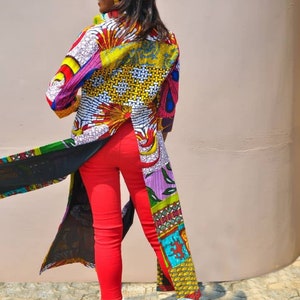 May include: A woman wearing a long, colourful patterned shirt with a black sheer overlay and red trousers. The shirt has a split in the back and is made of different fabrics with various patterns. The woman is standing in front of a light pink wall.