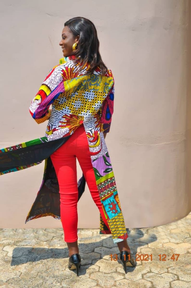 May include: A woman wearing a long, colourful patterned shirt with a black sheer overlay and red trousers. The shirt has a split in the back and is made of different fabrics with various patterns. The woman is standing in front of a light pink wall.