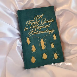 May include: A Field Guide to Magical Entomology book with a dark teal cover and gold lettering and insect illustrations. The book is resting on a white satin surface. The text "BEE SPLENDID" is at the bottom.