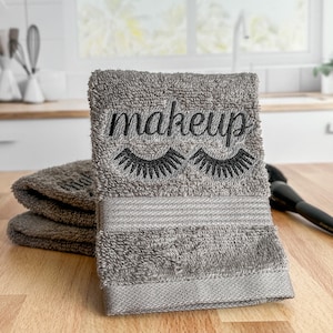 May include: Gray hand towel with the word "makeup" embroidered in black with eyelashes above the word.