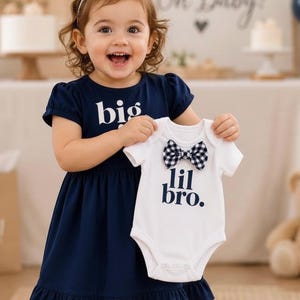 May include: A navy blue dress with short sleeves and the text big printed in white, paired with a white short-sleeved bodysuit featuring the text lil bro. in navy blue and a black and white checkered bow tie attached at the neckline.