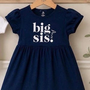 May include: A navy blue short-sleeved dress with a gathered waist and a ruffled hem. The front features the white text "big sis" with the name "Ava" printed vertically in white lettering next to the word "sis" on the chest area.
