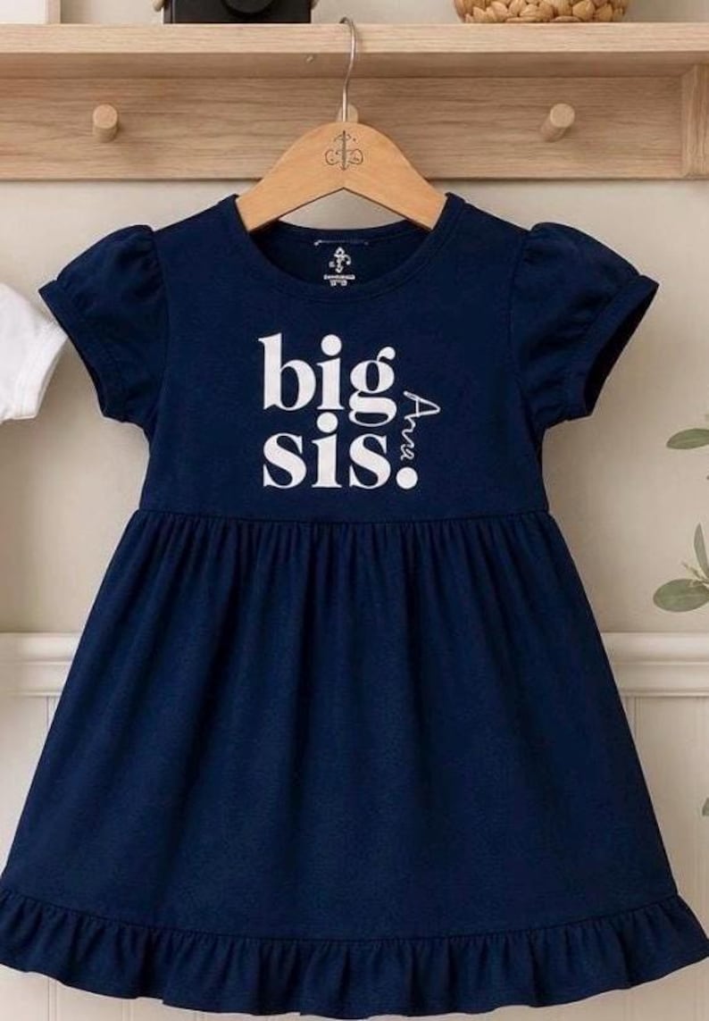 May include: A navy blue short-sleeved dress with a gathered waist and a ruffled hem. The front features the white text "big sis" with the name "Ava" printed vertically in white lettering next to the word "sis" on the chest area.