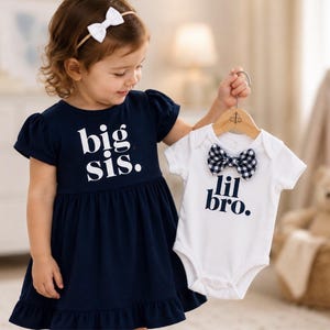 May include: A navy blue dress with short puff sleeves and a ruffled hem featuring the white text big sis. and a white short-sleeved bodysuit with a black and white gingham bow tie and the dark blue text lil bro. on a wooden hanger.