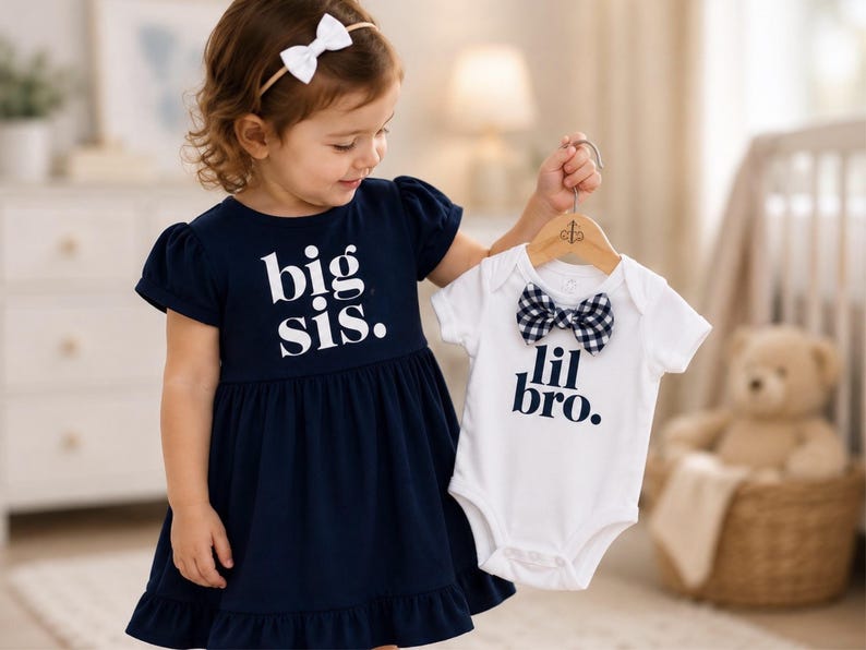 May include: A navy blue dress with short puff sleeves and a ruffled hem featuring the white text big sis. and a white short-sleeved bodysuit with a black and white gingham bow tie and the dark blue text lil bro. on a wooden hanger.