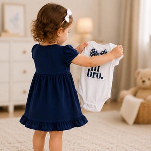 May include: A navy blue short-sleeved dress with a ruffled hem and a white baby bodysuit featuring a navy blue gingham bow tie and the text "lil bro." printed in navy blue letters on the front, held up by a child wearing a white hair bow.