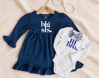 Puede incluir: Un vestido azul marino de manga larga con dobladillo con volantes y el texto blanco big sis. junto a un body blanco de manga larga con pajarita azul marino y el texto azul marino lil bro. El vestido cuelga de una percha de madera.