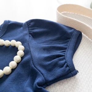 May include: A dark blue short-sleeved shirt featuring gathered puff sleeves and a round neckline. A strand of white faux pearl beads is draped across the front of the fabric. The garment is made of a solid-colored knit material with visible stitching.