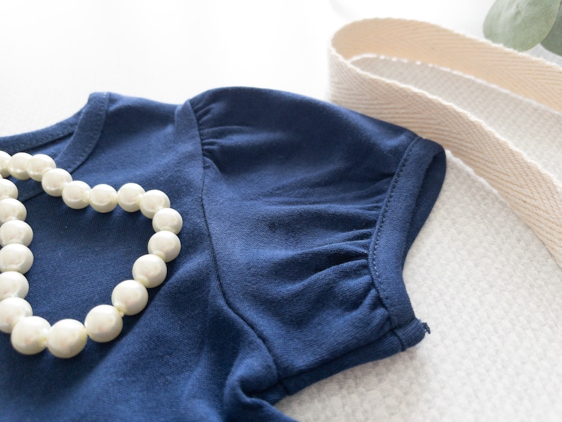 May include: A dark blue short-sleeved shirt featuring gathered puff sleeves and a round neckline. A strand of white faux pearl beads is draped across the front of the fabric. The garment is made of a solid-colored knit material with visible stitching.
