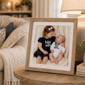 May include: A square wooden picture frame containing a photograph of a young child wearing a dark blue dress with the text big sis and a baby wearing a white onesie with the text lil bro. The frame has a light brown finish and a white mat border.