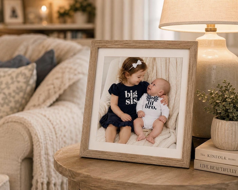 May include: A square wooden picture frame containing a photograph of a young child wearing a dark blue dress with the text big sis and a baby wearing a white onesie with the text lil bro. The frame has a light brown finish and a white mat border.