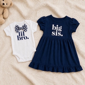 May include: A white short-sleeved bodysuit with a navy and white gingham bow tie and the text "lil bro." in navy, alongside a navy blue short-sleeved dress with a ruffled hem and the text "big sis." printed in white on the chest.