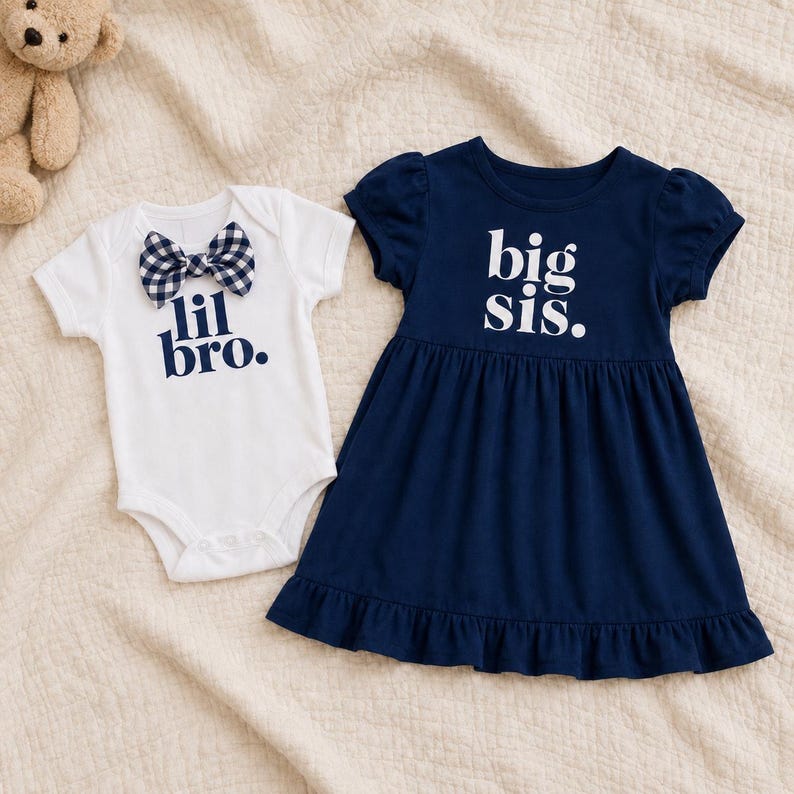 May include: A white short-sleeved bodysuit with a navy and white gingham bow tie and the text "lil bro." in navy, alongside a navy blue short-sleeved dress with a ruffled hem and the text "big sis." printed in white on the chest.