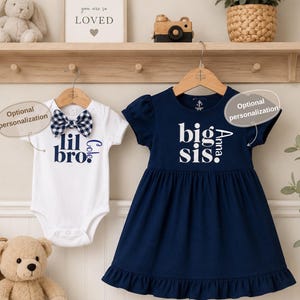 May include: A white short-sleeved bodysuit with a navy and white gingham bow tie and the text "lil bro" in navy blue, paired with a navy blue short-sleeved dress featuring a ruffled hem and the white text "big sis" printed on the front.