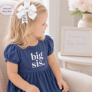 May include: A navy blue dress with short puff sleeves and the white text big sis. printed on the chest. A white hair bow with the text big sis. is attached to the hair. The bow is labeled as an optional hair bow in the image text.