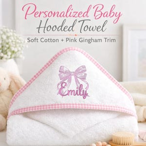 May include: White baby hooded towel with pink gingham trim and a pink embroidered bow and name. The text reads "Personalized Baby Hooded Towel" and "Soft Cotton + Pink Gingham Trim". A plush bunny and a brush are in the background.