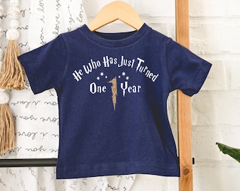 May include: Navy blue short-sleeved t-shirt with the text "He Who Has Just Turned One Year" in white and gold. The design includes a lightning bolt and stars. The shirt is displayed on a wooden hanger.