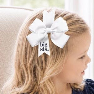 May include: A white hair bow made of smooth ribbon with multiple loops and a central knot. One of the downward-hanging ribbon tails features the text "big sis." printed in a dark, bold, sans-serif font, centered on the white fabric surface.