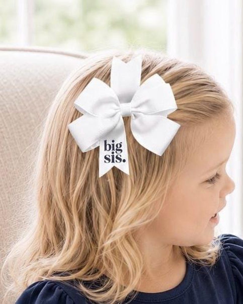 May include: A white hair bow made of smooth ribbon with multiple loops and a central knot. One of the downward-hanging ribbon tails features the text "big sis." printed in a dark, bold, sans-serif font, centered on the white fabric surface.