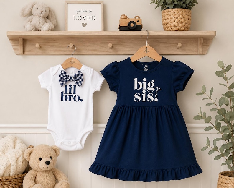 May include: A white short-sleeved bodysuit with a navy blue gingham bow tie and the text 'lil bro.' paired with a navy blue short-sleeved dress featuring a ruffled hem and the white text 'big sis.' printed on the front of the bodice.