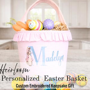 May include: Pink gingham Easter basket with a matching handle and ruffle trim. The basket is personalized with a name in blue script and a bunny illustration. It contains decorative eggs, carrots, and straw.