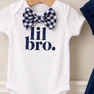May include: A white short-sleeved bodysuit featuring a navy blue and white gingham bow tie at the neckline. The text "lil bro." is printed in navy blue on the front of the garment. The bodysuit is displayed on a wooden hanger.