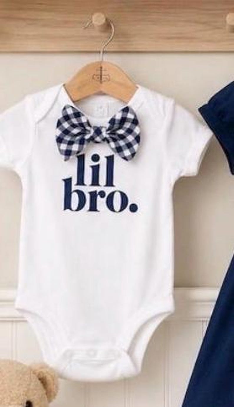 May include: A white short-sleeved bodysuit featuring a navy blue and white gingham bow tie at the neckline. The text "lil bro." is printed in navy blue on the front of the garment. The bodysuit is displayed on a wooden hanger.