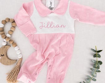 May include: A long-sleeved baby romper in light pink and white featuring a Peter Pan collar and the name Jillian embroidered in pink script across the chest. A white tag at the neckline displays the text NB and a small logo icon.