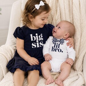 May include: A navy blue dress with short puff sleeves and the white text big sis. paired with a white long-sleeve bodysuit featuring the navy blue text lil bro. and a navy and white gingham bowtie. The dress has a ruffled hemline.