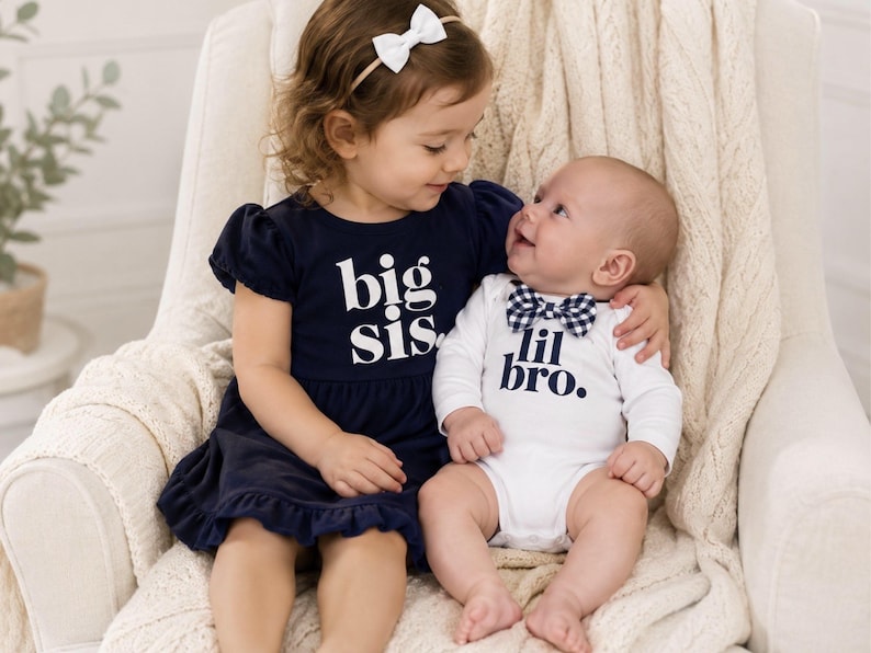 May include: A navy blue dress with short puff sleeves and the white text big sis. paired with a white long-sleeve bodysuit featuring the navy blue text lil bro. and a navy and white gingham bowtie. The dress has a ruffled hemline.