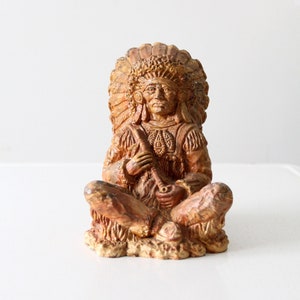 May include: A vintage figurine of a Native American chief in a seated position, wearing a feathered headdress. The statue is a warm brown color with intricate details, holding a pipe. The figurine is likely made of resin or a similar material.