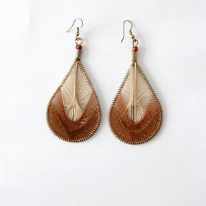 May include: A pair of teardrop-shaped earrings. The earrings feature a woven design with a gradient of colors, transitioning from beige to brown. The earrings have a metal hook and a small bead at the top. The background is plain white.