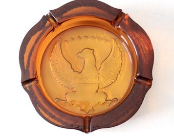 May include: Amber-colored glass ashtray with a brown rim. The ashtray features a detailed embossed design of an eagle with outstretched wings, surrounded by stars. The ashtray has four notches for resting cigarettes.