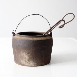 Op de afbeelding: Een vintage gietijzeren pot met een draadhandvat en een draadbeugel. De pot is roestig en heeft een versleten oppervlak.