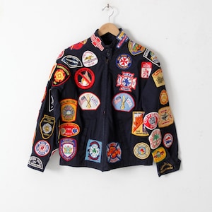 Puede incluir: Una chaqueta azul marino con una variedad de parches bordados, muchos de los cuales están relacionados con el departamento de bomberos. Los parches son de varios colores y diseños, incluyendo rojo, amarillo, azul y blanco.